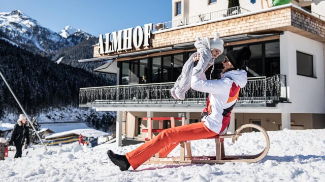 Mutter hebt Kind im Schnee vor Almhotel hoch